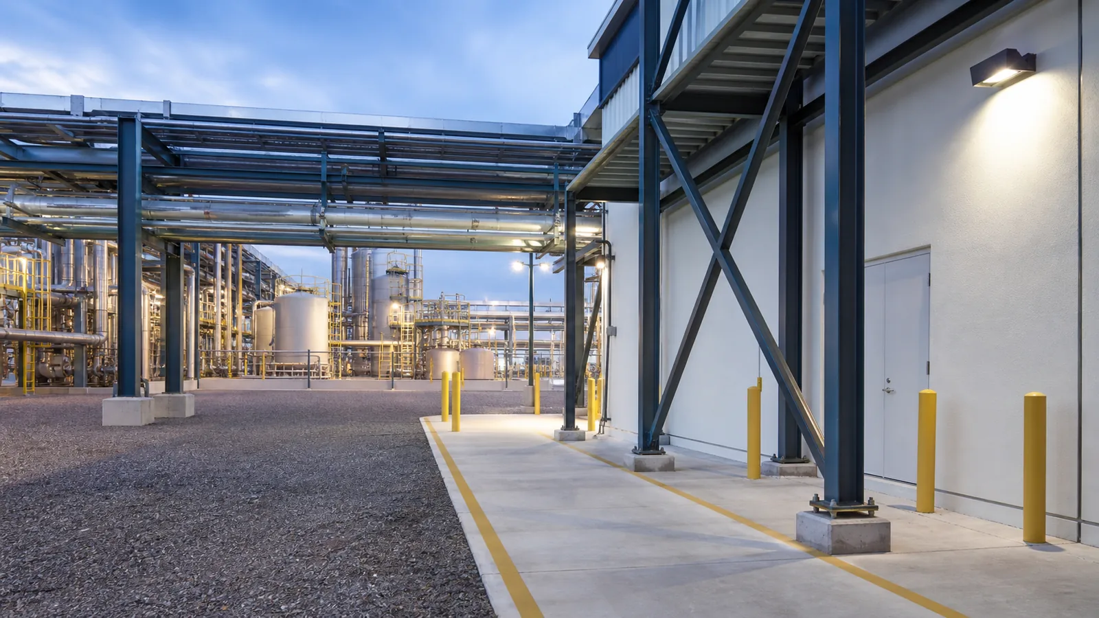 Chemical manufacturing facility with process piping in the distance, an isolated maintenance building, freshly painted structural steel, and controlled access paths